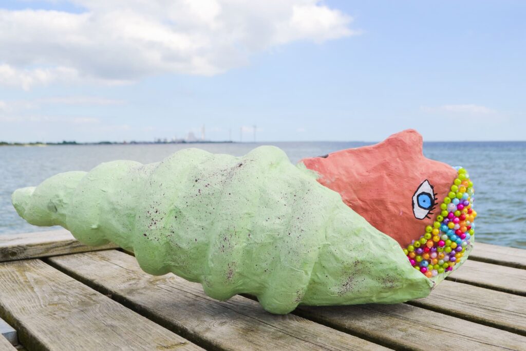 En grøn konkylie lavet af papmaché har fået perlebadehat på og slikker sol på en badebro